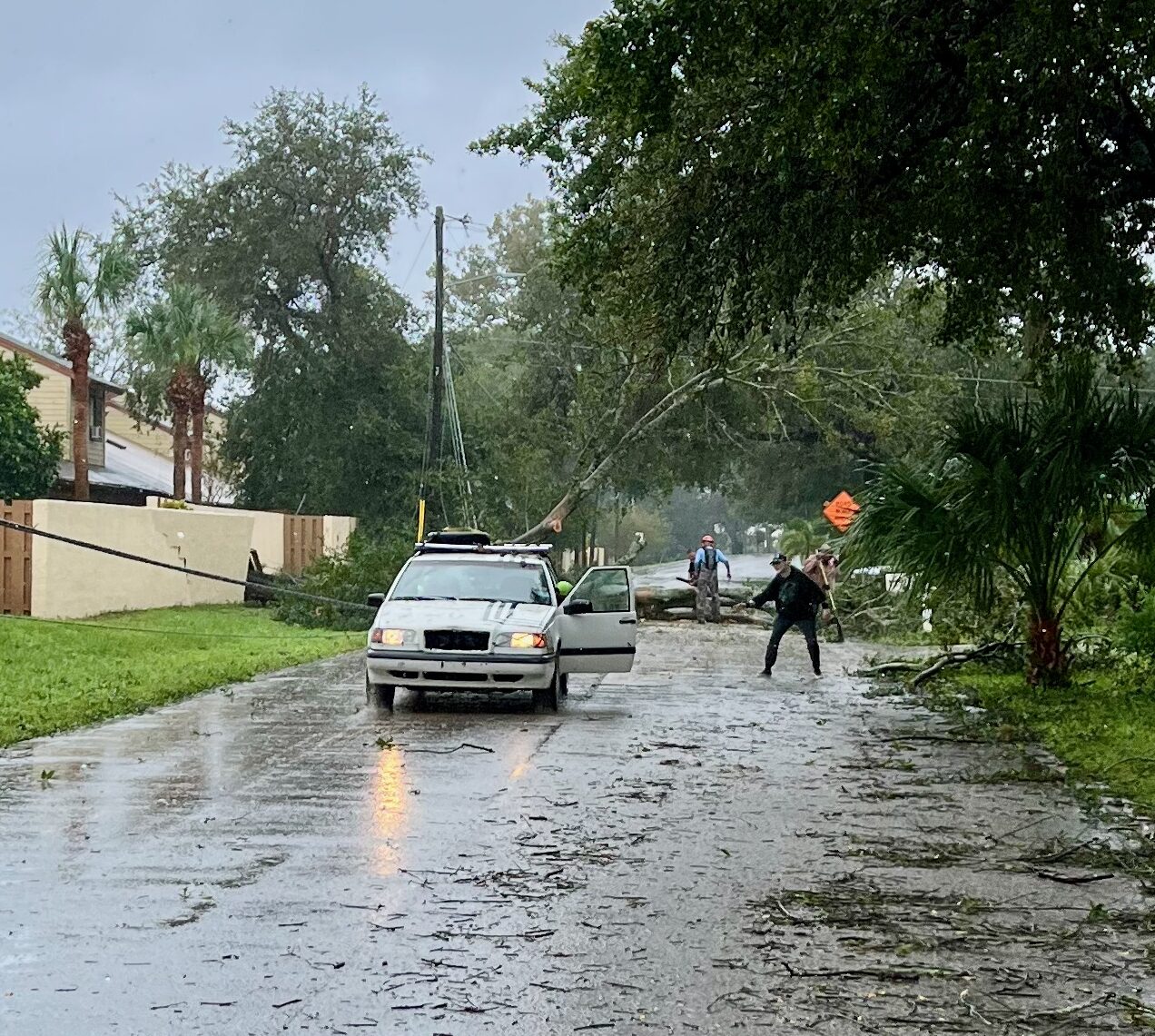 A residential street on a rainy day after a Hurricane. A large tree fallen and blocks the entire street. A power line is down on the side of the street. Neighbors with chain saws are quickly cutting through the tree to clear it from the road. A white Volvo station wagon is parked in front of the tree to pull away larger debris. A neighbor dressed in black pants and a black jacket and hat is directing the car as it backs up to a large tree limb.